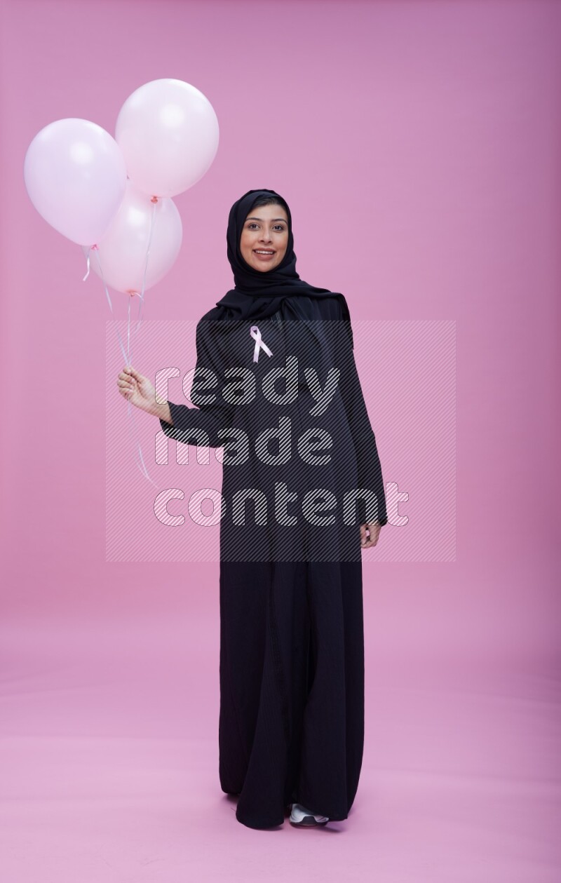 Saudi woman wearing pink ribbon on Abaya standing holding balloons on plain pink background