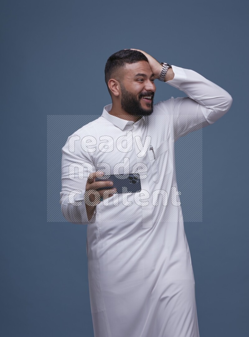 A man Playing a Game on his Smartphone  on Blue Background wearing Saudi Thob
