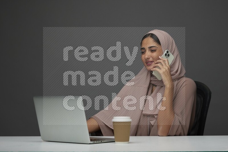 A Saudi woman Setting on her desk
 calling  on a Gray Background wearing Brown Abaya with Hijab