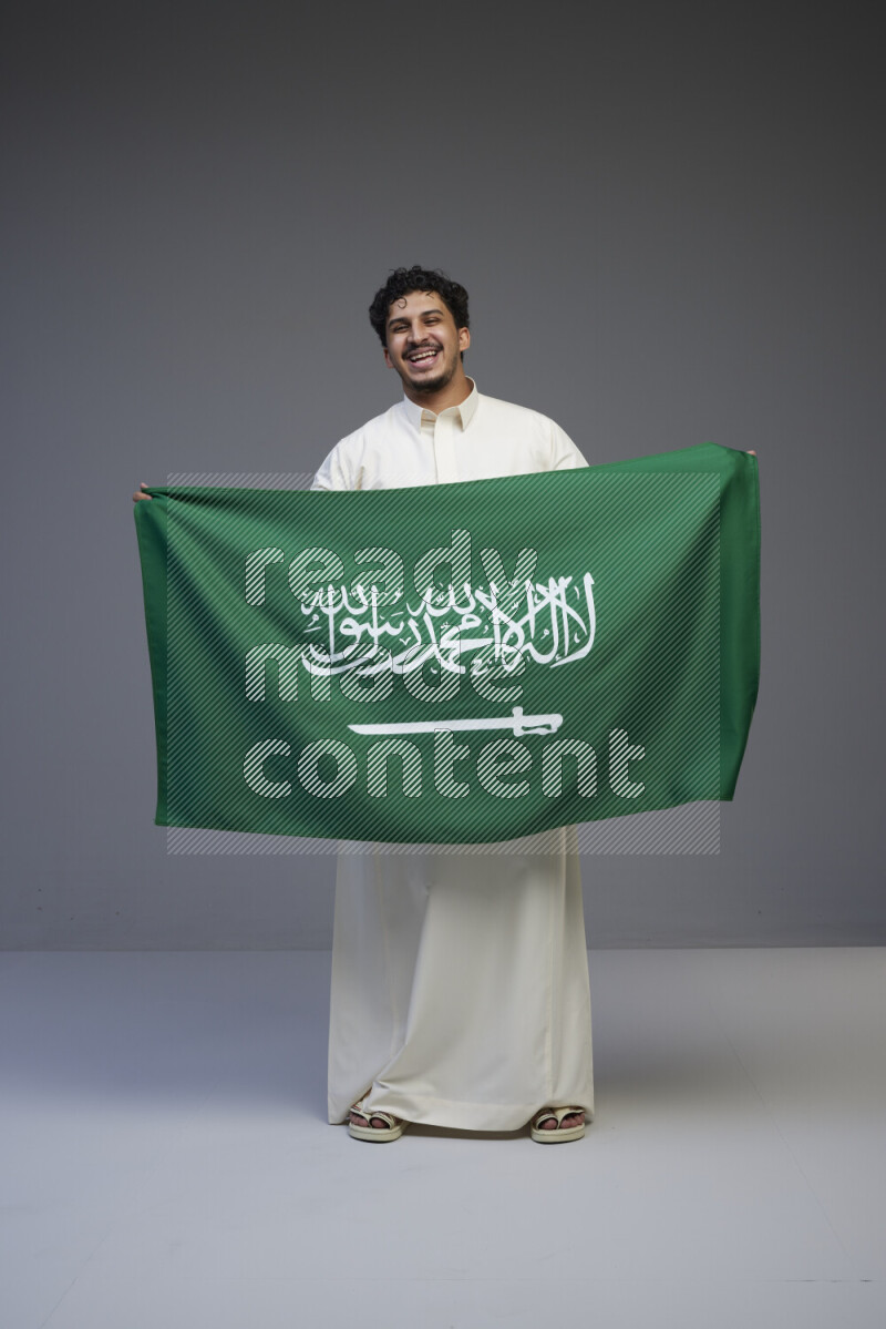 A Saudi man standing wearing thob holding big Saudi flag on gray background