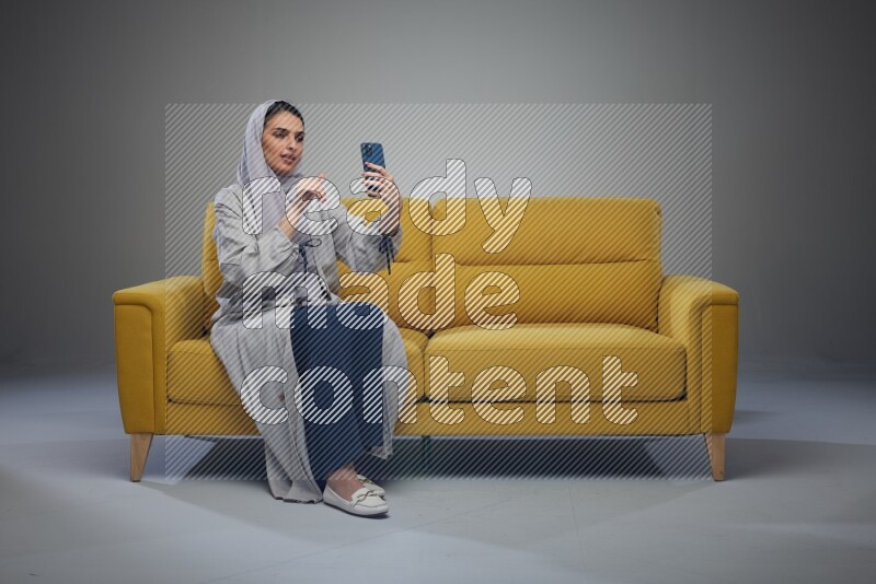 A Saudi woman wearing a light gray Abaya and head scarf sitting on a yellow sofa and making a video call eye level on a grey background
