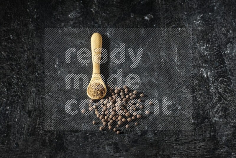 A wooden spoon full of allspice powder and whole balls spreaded on a textured black flooring