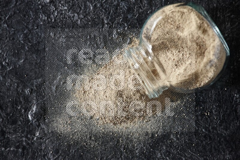 A flipped herbal glass jar full of white pepper powder with spilled powder on textured black flooring