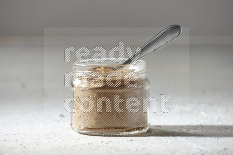 A glass jar and a metal spoon full of cumin powder on textured white flooring