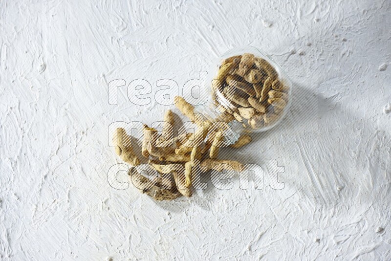 A flipped glass spice jar full of dried turmeric fingers and fell out it on textured white flooring