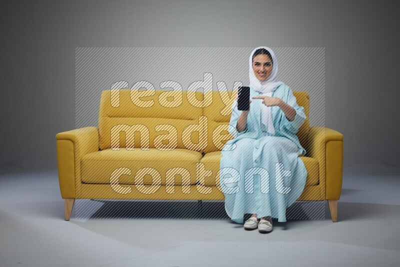 A Saudi woman wearing a light blue Abaya and white head scarf sitting on a yellow sofa and showing her phone's screen vertically and horizontally eye level on a grey background