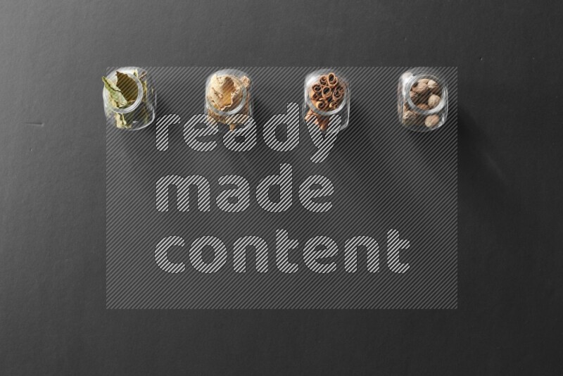 4 glass spice jars full of laurel bay leaves, ginger, cinnamon and nutmeg on black flooring