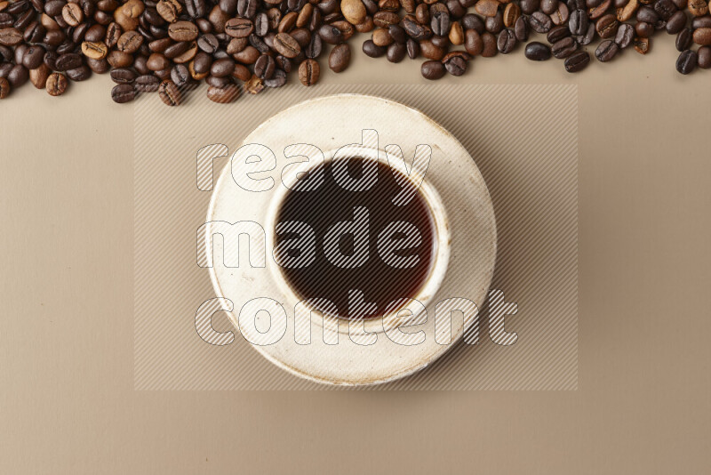 A beige pottery cup of coffee surrounded by roasted coffee beans on beige background