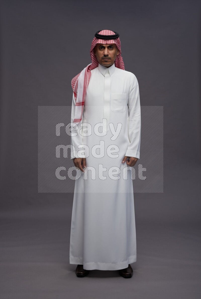 Saudi man wearing thob and shomag standing interacting with the camera on gray background