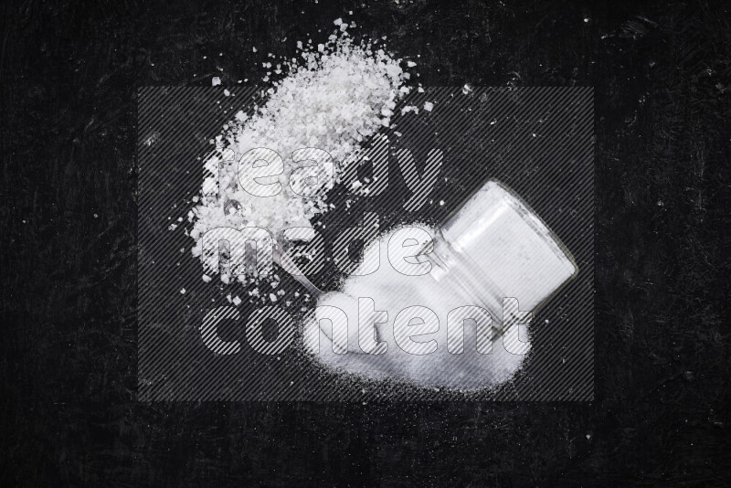 A glass jar full of table salt with some sea salt crystals beside it on a black background