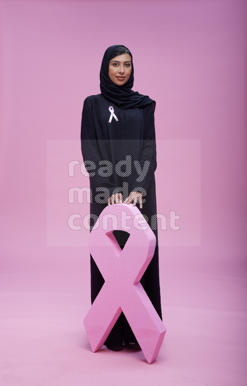 Saudi woman wearing pink ribbon on abaya standing holding awareness ribbon on plain pink background