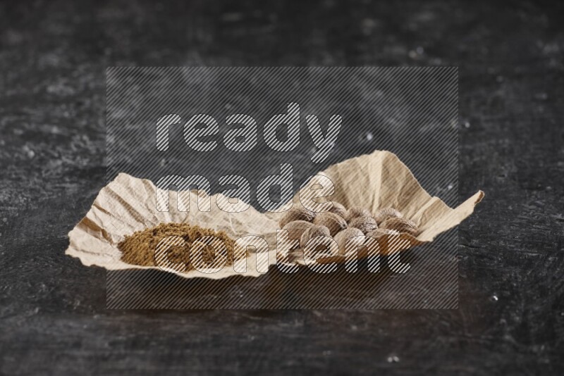 2 crumpled piece of paper full of nutmeg seeds and powder on a textured black flooring