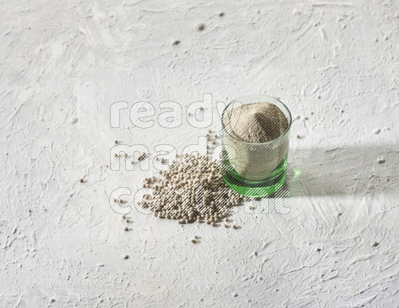A green glass cup full of white pepper powder with white pepper beads on textured white flooring