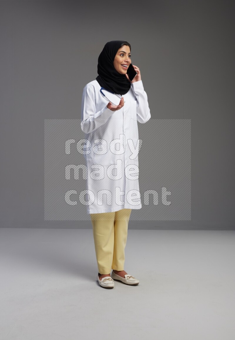 Saudi woman wearing lab coat with stethoscope standing talking on phone on Gray background