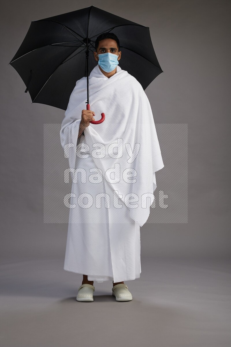 A man wearing Ehram and mask Standing holding umbrella on gray background on gray background