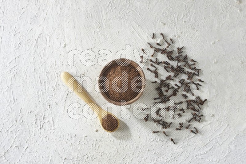 A wooden bowl and wooden spoon full of cloves powder with cloves spread on textured white flooring