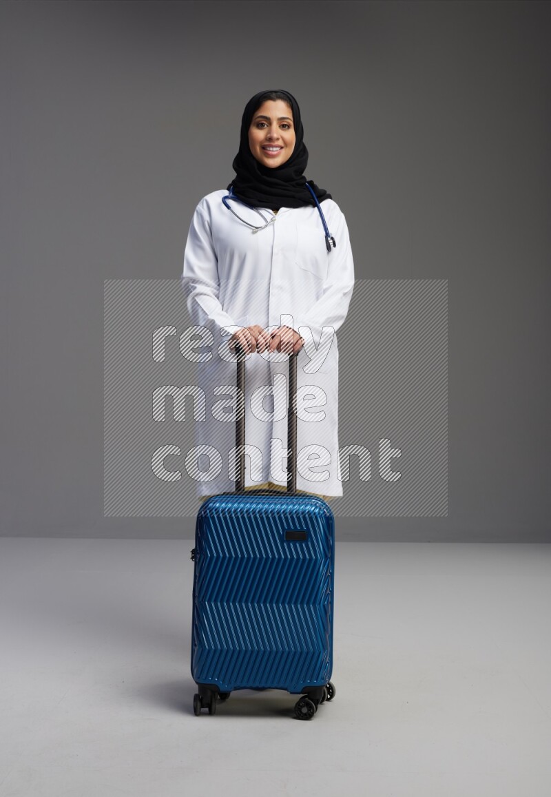 Saudi woman wearing lab coat with stethoscope standing holding Travel bag on Gray background
