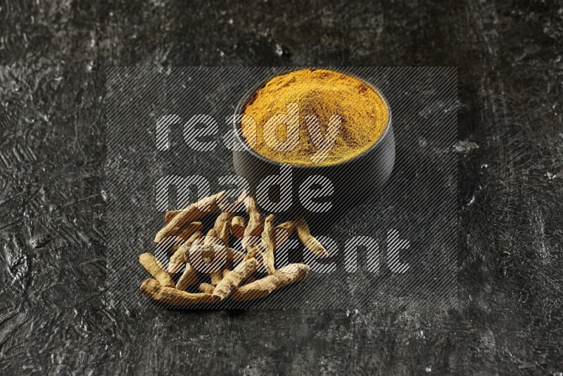 A black pottery bowl full of turmeric powder and dried turmeric whole fingers next of it on textured black flooring