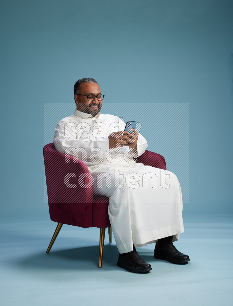 Saudi Man without shimag sitting on chair texting on phone on blue background