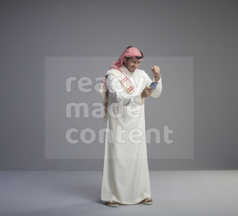 A  Saudi man standing wearing thob and red shomag texting on phone on gray background