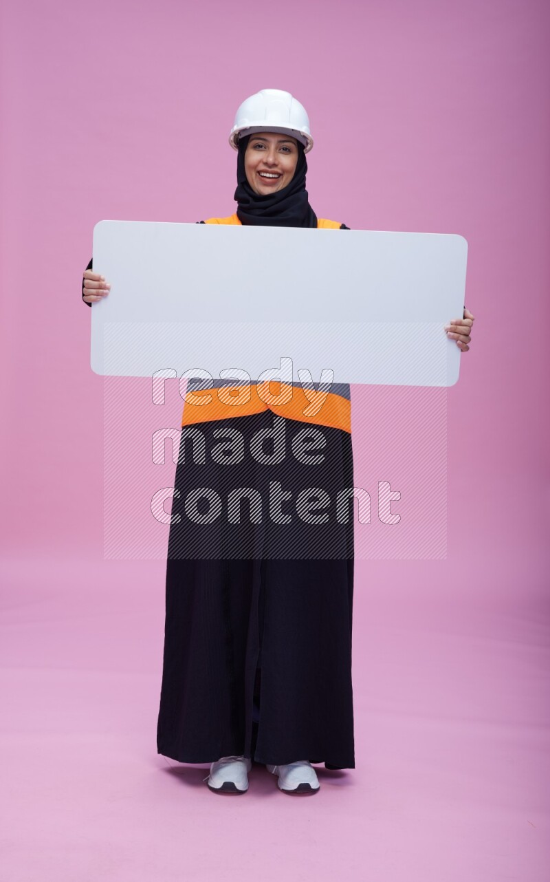 Saudi woman wearing Abaya with engineer vest and helmet standing holding board on pink background