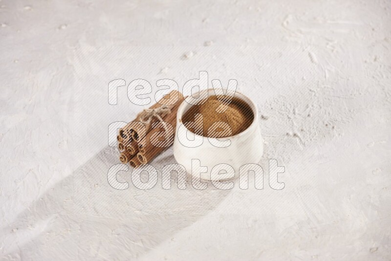 Cinnamon sticks stacked and bounded beside a beige bowl full of cinnamon powder on white background