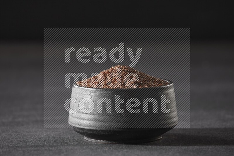 A black pottery bowl full of flaxseeds on a black flooring