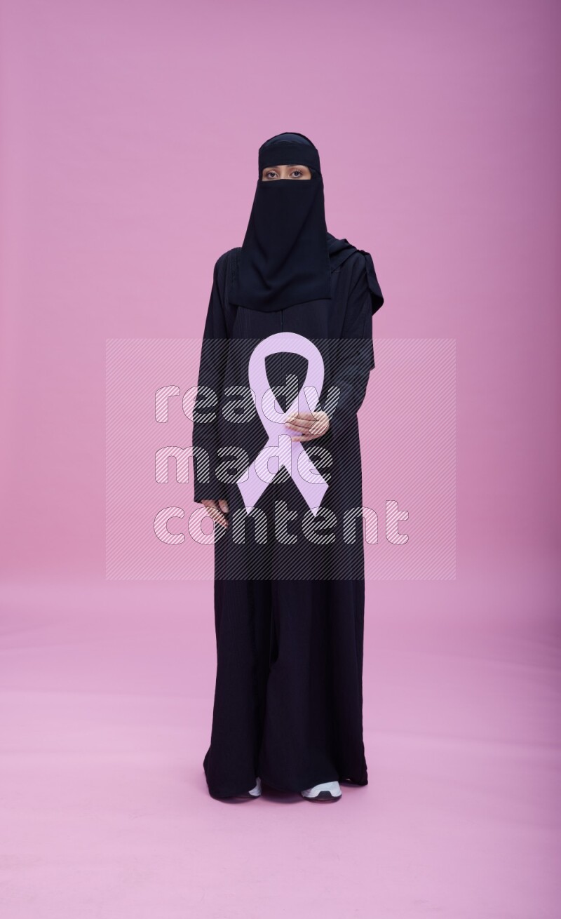 Saudi woman wearing abaya and niqab standing holding awareness ribbon on pink background
