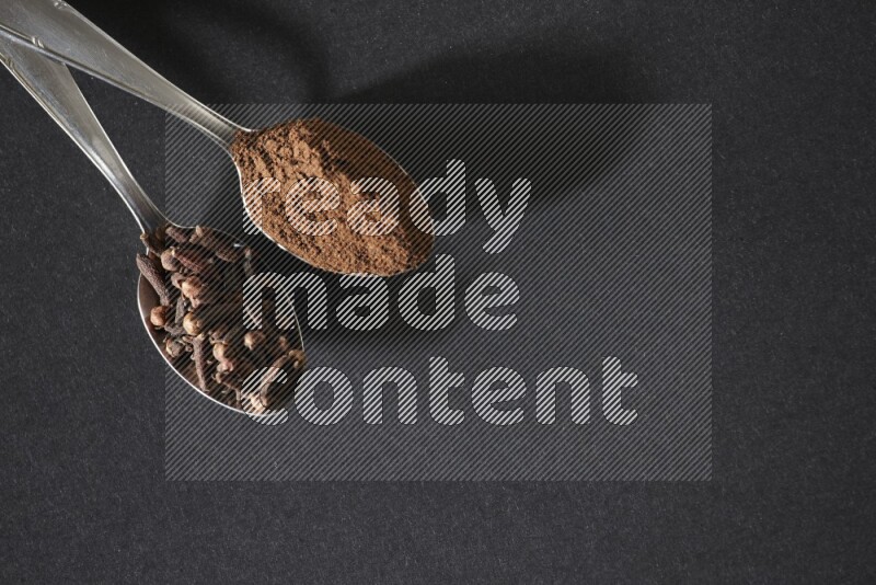 2 metal spoons, one full of cloves grains and the other full of cloves powder on a black flooring
