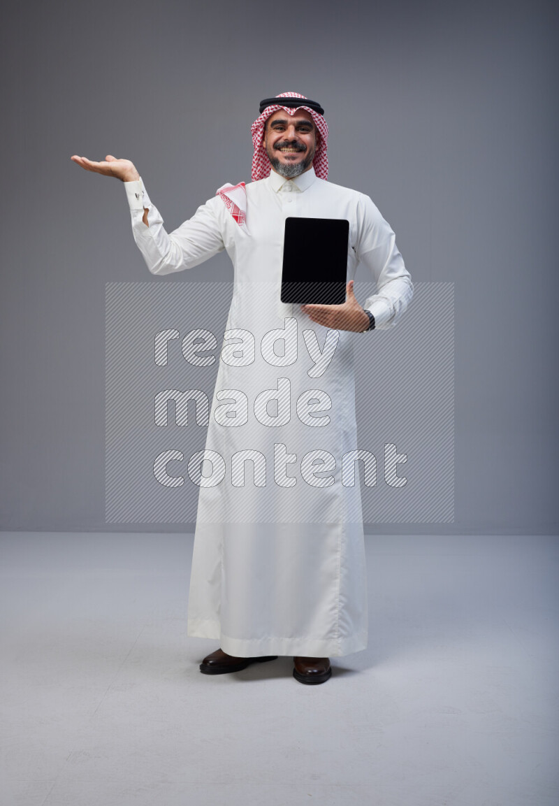 Saudi man Wearing Thob and red Shomag standing showing tablet to camera on Gray background