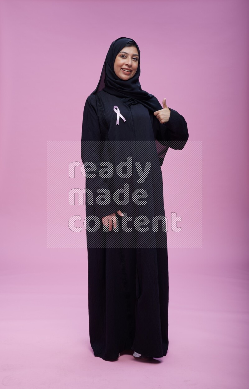 Saudi woman wearing pink ribbon on Abaya standing interacting with the camera on pink background