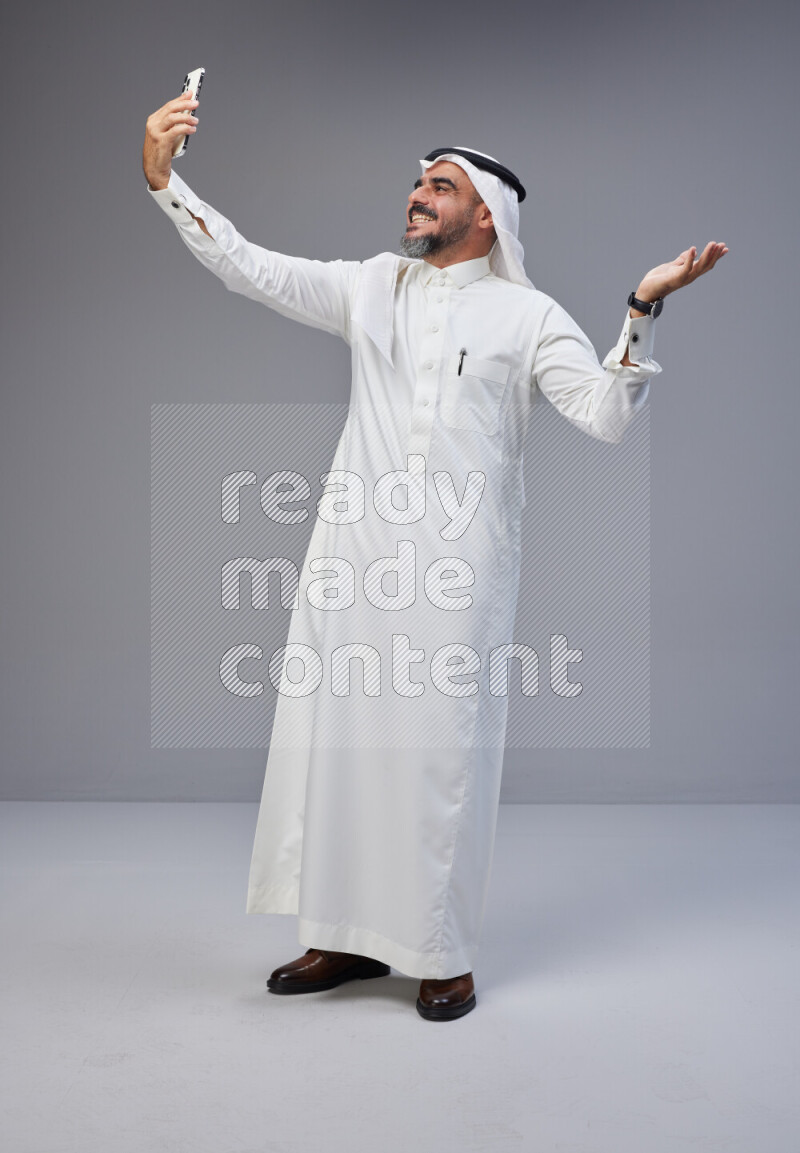 Saudi man Wearing Thob and white Shomag standing taking selfie on Gray background