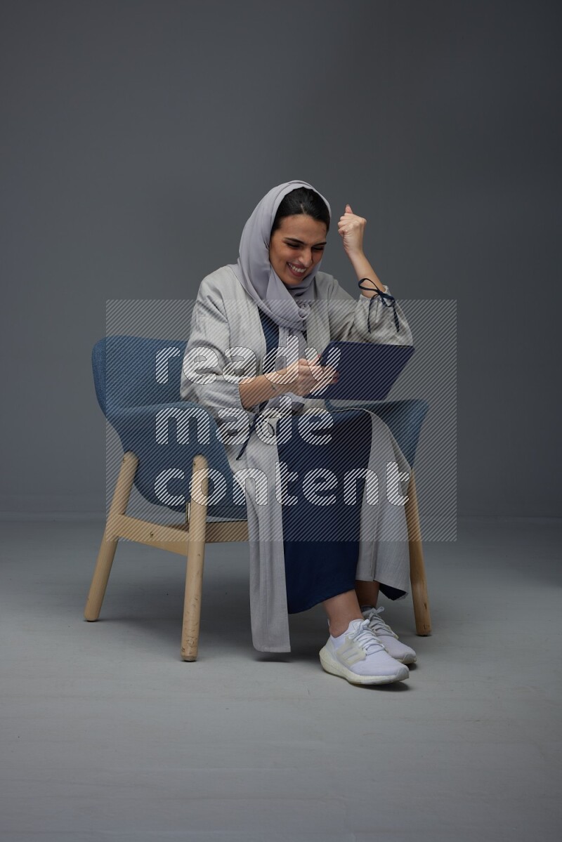 A Saudi female wearing a light gray Abaya and head scarf sitting on a dark grey chair while holding her iPad on a grey background