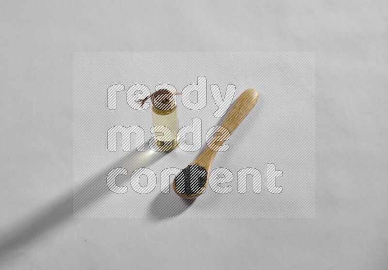 A wooden spoon full of black seeds with a glass bottle of black seeds oil on a white flooring