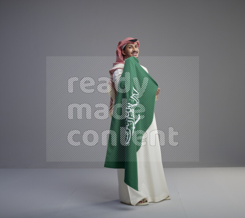 رجل سعودي يرتدي ثوب ابيض وشماغ احمر ويحمل علم كبير للسعودية