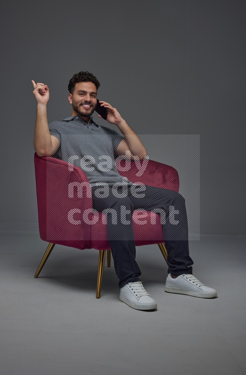 A man wearing casual talking in the phone and making multi hand gestures while sitting on a burgundy chair different angles eye level on a gray background