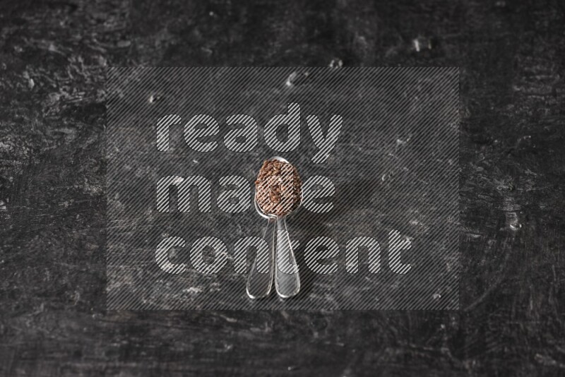 2 metal spoons full of flaxseeds on a textured black flooring