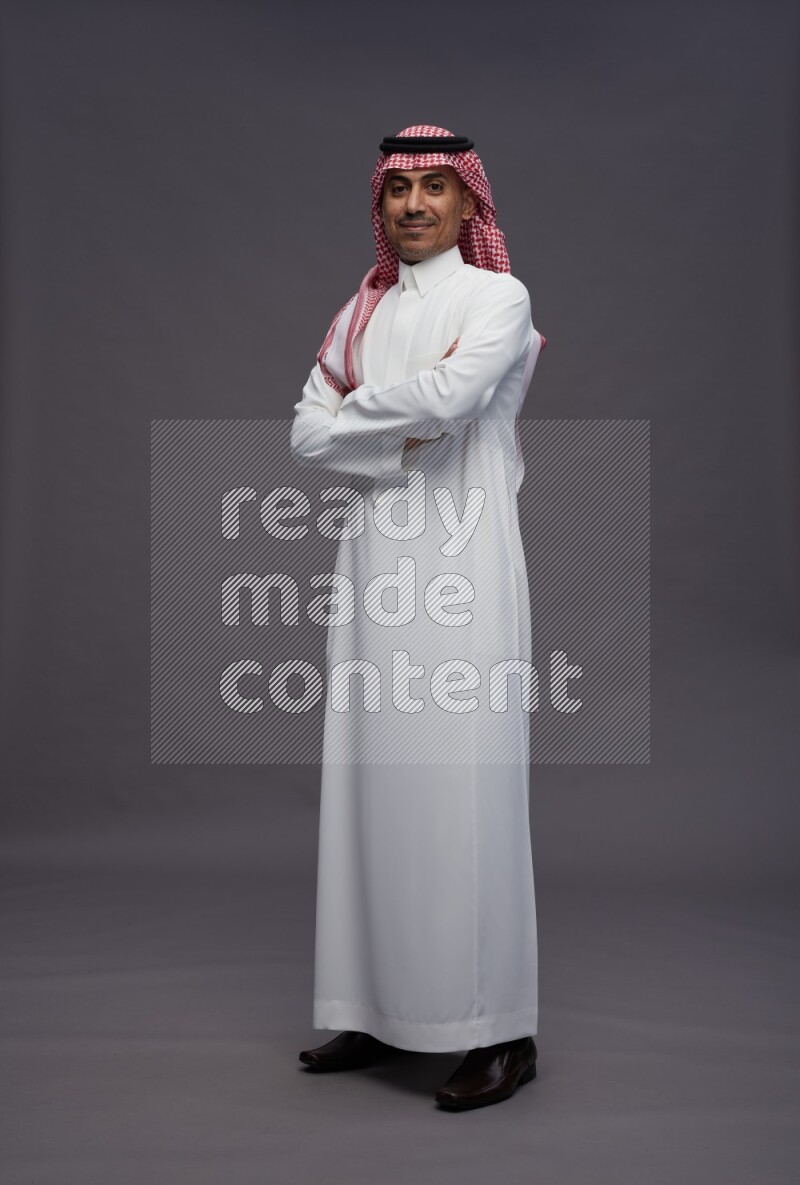 Saudi man wearing thob and shomag standing with crossed arms on gray background