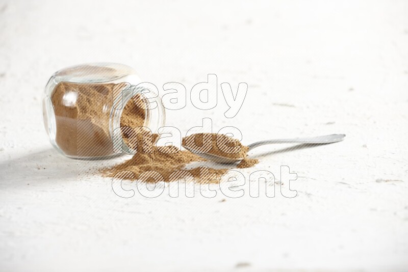 Flipped herbs glass jar full of cinnamon powder with a metal spoon full of powder on a textured white background