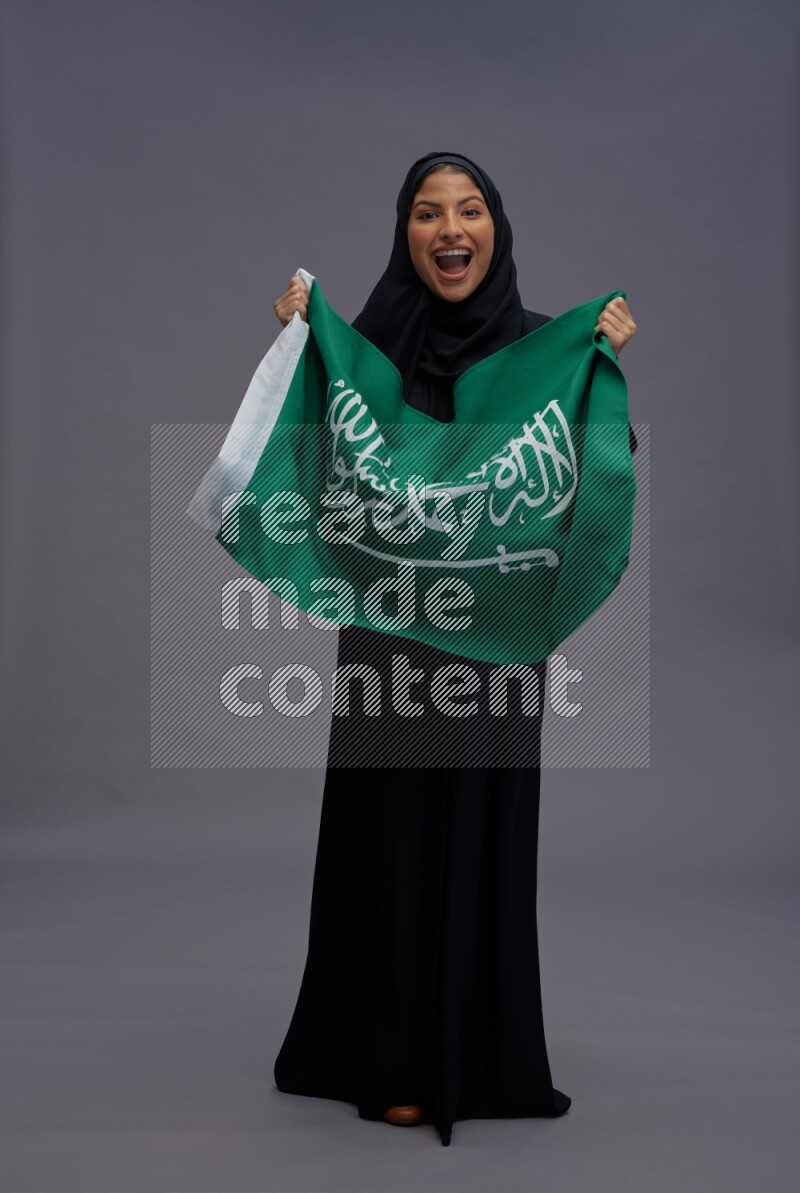Saudi woman wearing Abaya standing holding Saudi flag on gray background