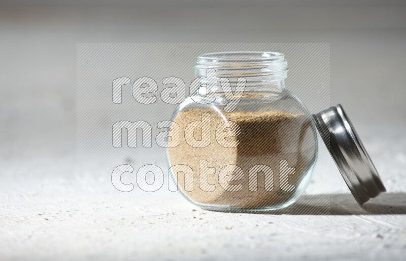 A glass spice jar full of cumin powder on textured white flooring