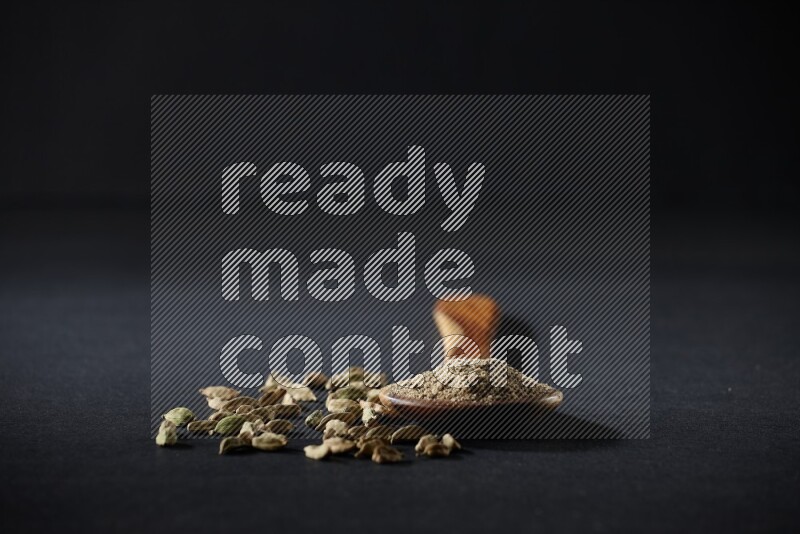 A wooden ladle full of cardamom powder and cardamom seeds beside it on black flooring