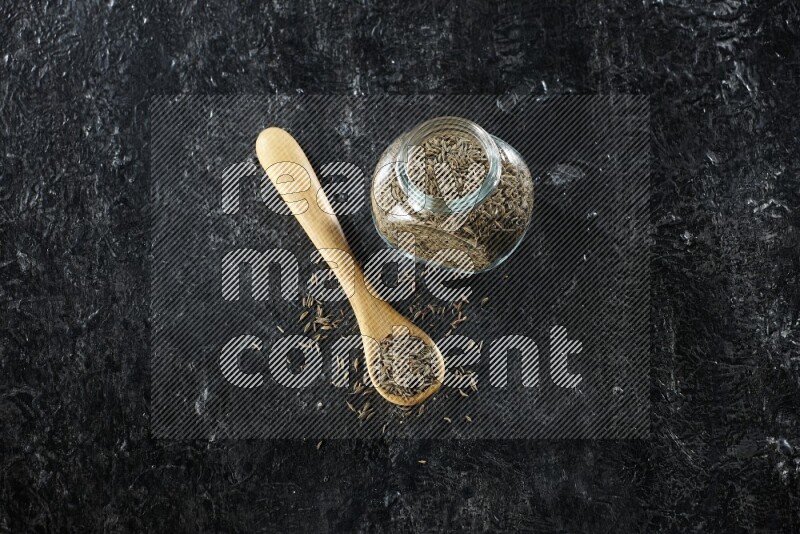 A glass spice jar and a wooden spoon full of cumin seeds on a textured black flooring
