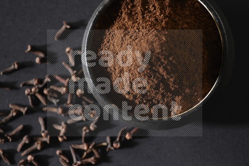 وعاء فخاري أسود ممتلئ ببودرة القرنفل وحبوب القرنفل على خلفية سوداء