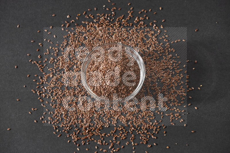 A glass bowl full of flaxseeds surrounded by the seeds on a black flooring
