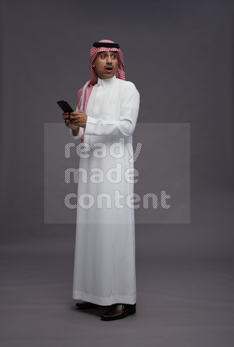 Saudi man wearing thob and shomag standing texting on phone on gray background
