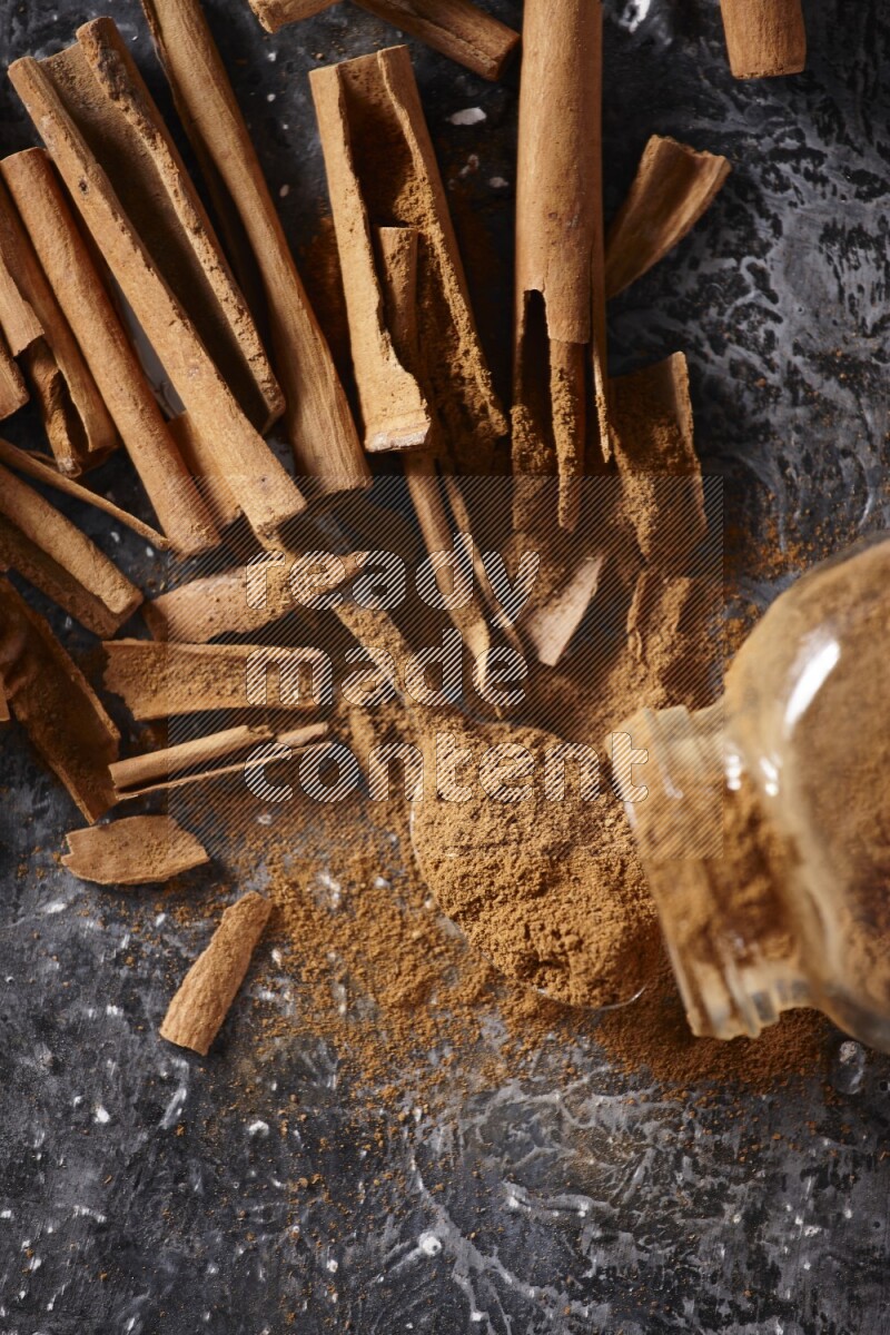 Herbal glass jar full cinnamon powder flipped and a metal spoon full of powder surrounded by cinnamon sticks on textured black background in different angles
