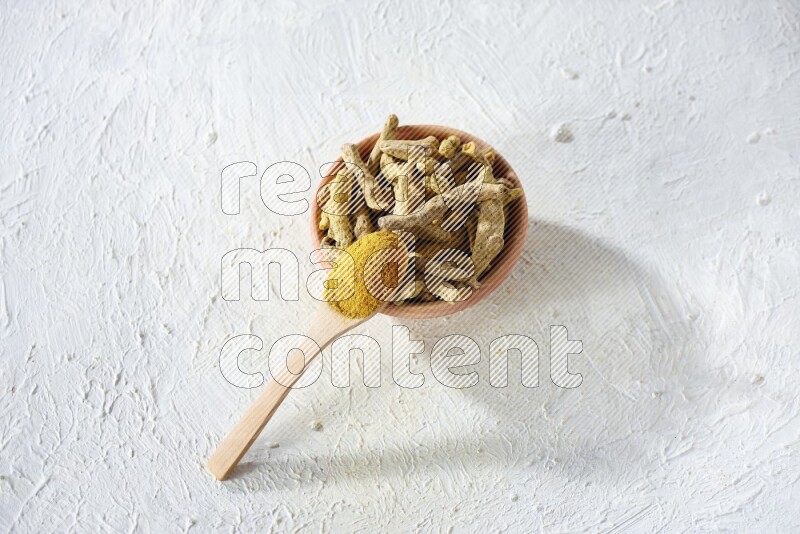 A wooden spoon full of turmeric powder above a wooden bowl full of dried turmeric whole fingers on a textured white flooring