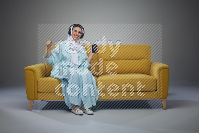 A Saudi woman wearing a light blue Abaya and a white head scarf sitting on a yellow sofa and holding an electronic while wearing headphone eye level on a grey background