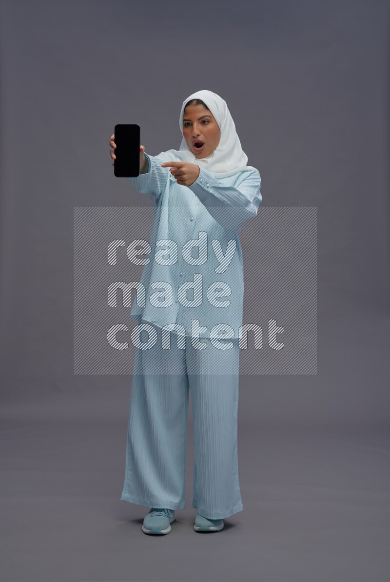 Saudi woman wearing hijab clothes standing showing phone to camera on gray background